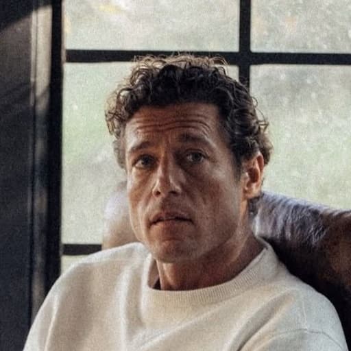 A curly-haired man in a white sweater seated indoors.
