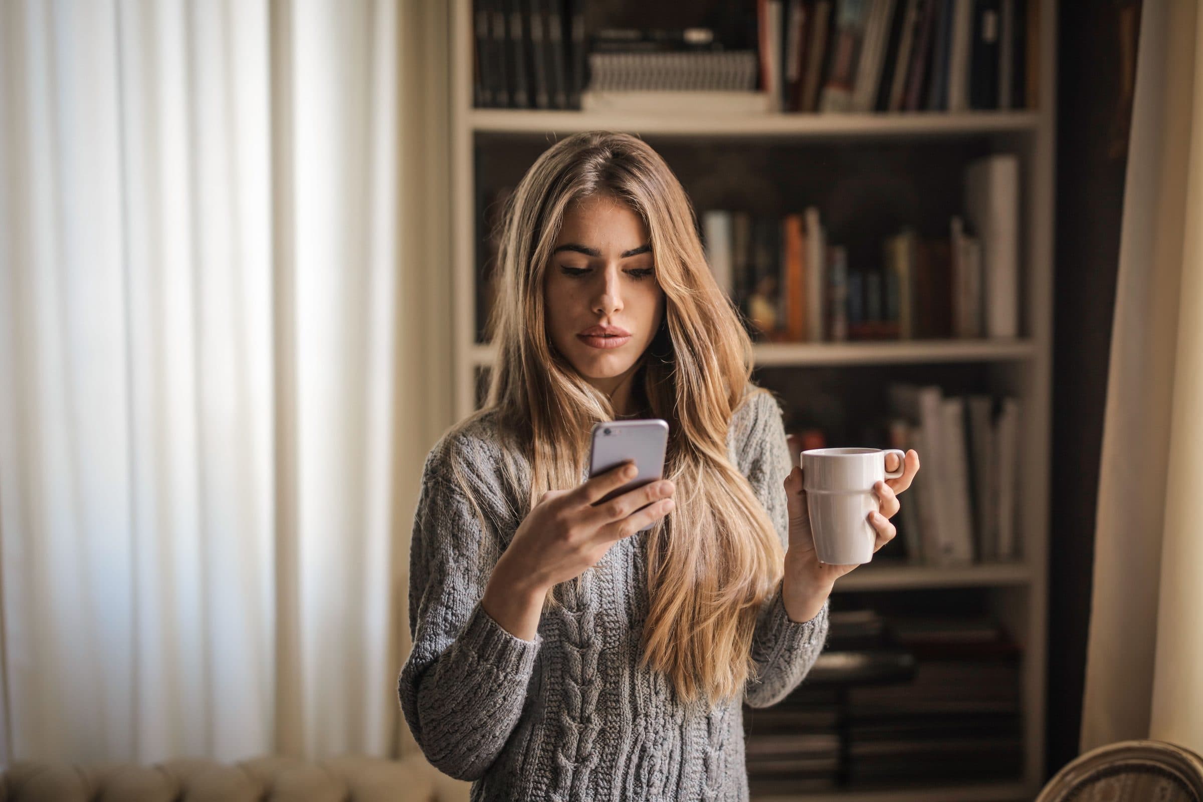 Woman with long highlighted hair in a grey cable-knit sweater looking unimpressed at her phone, holding a coffee mug