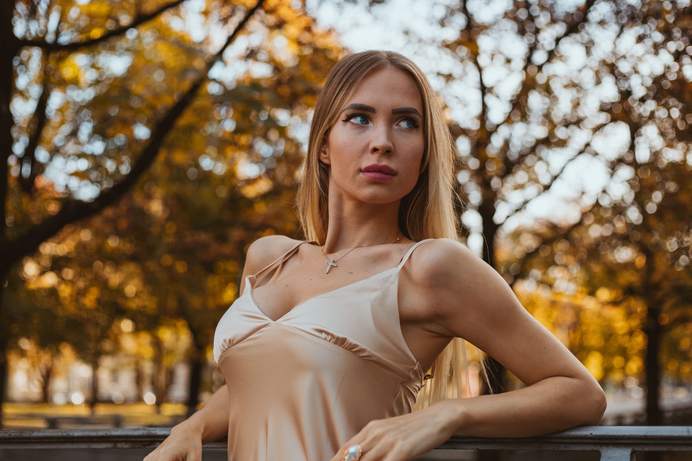 Woman with long blonde hair in a satin camisole leaning on a railing among autumn trees