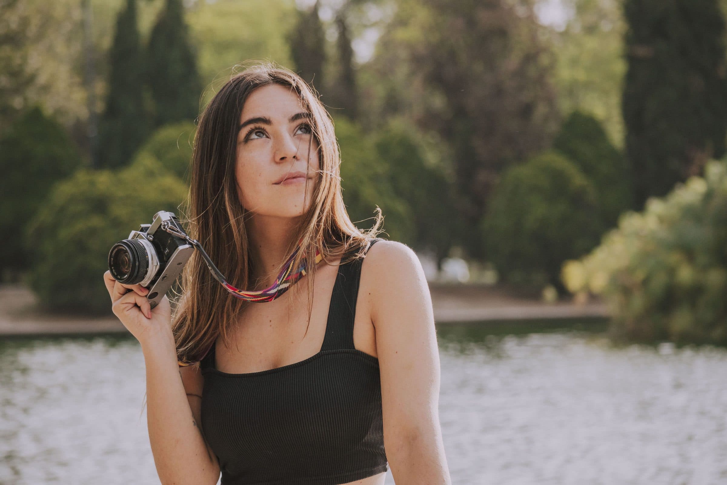 Woman with long brown hair in a black crop top holding a film camera by a pond in a park