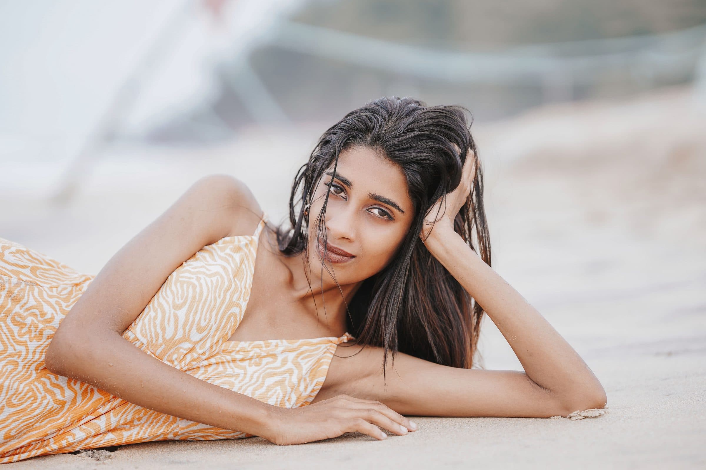 Woman with wet dark hair in a yellow patterned dress lying on sand, looking at the camera