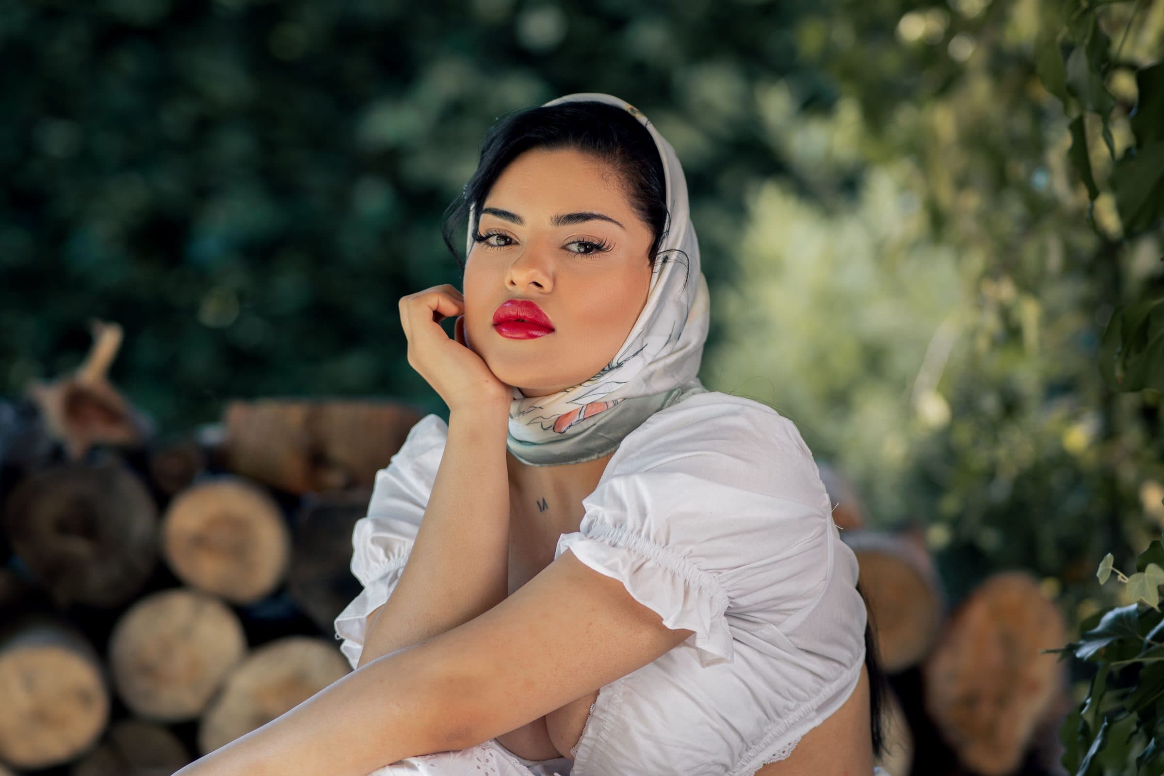 Woman in a silk headscarf and white blouse with red lips resting her cheek on her hand by stacked logs