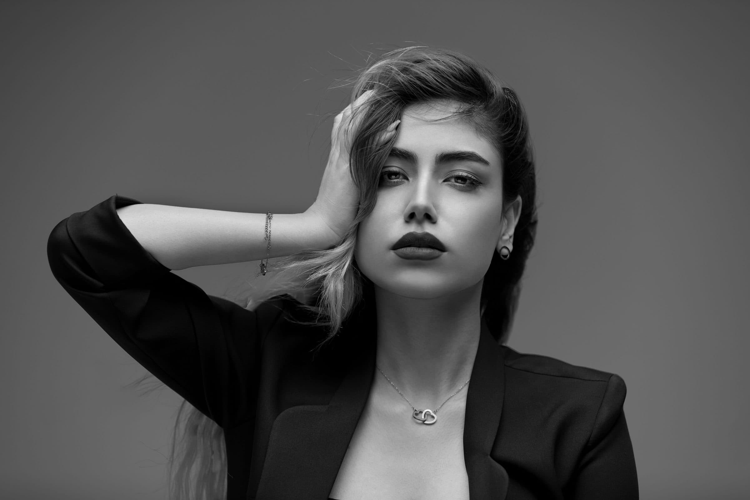 Black and white studio portrait of a woman with long hair pushed back by her hand, wearing a dark blazer and necklace