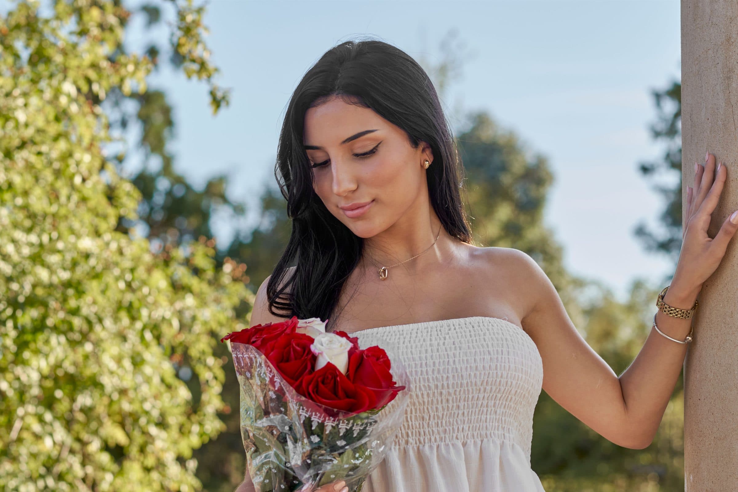 Woman with long dark hair in a white strapless top holding red roses and leaning on a pillar