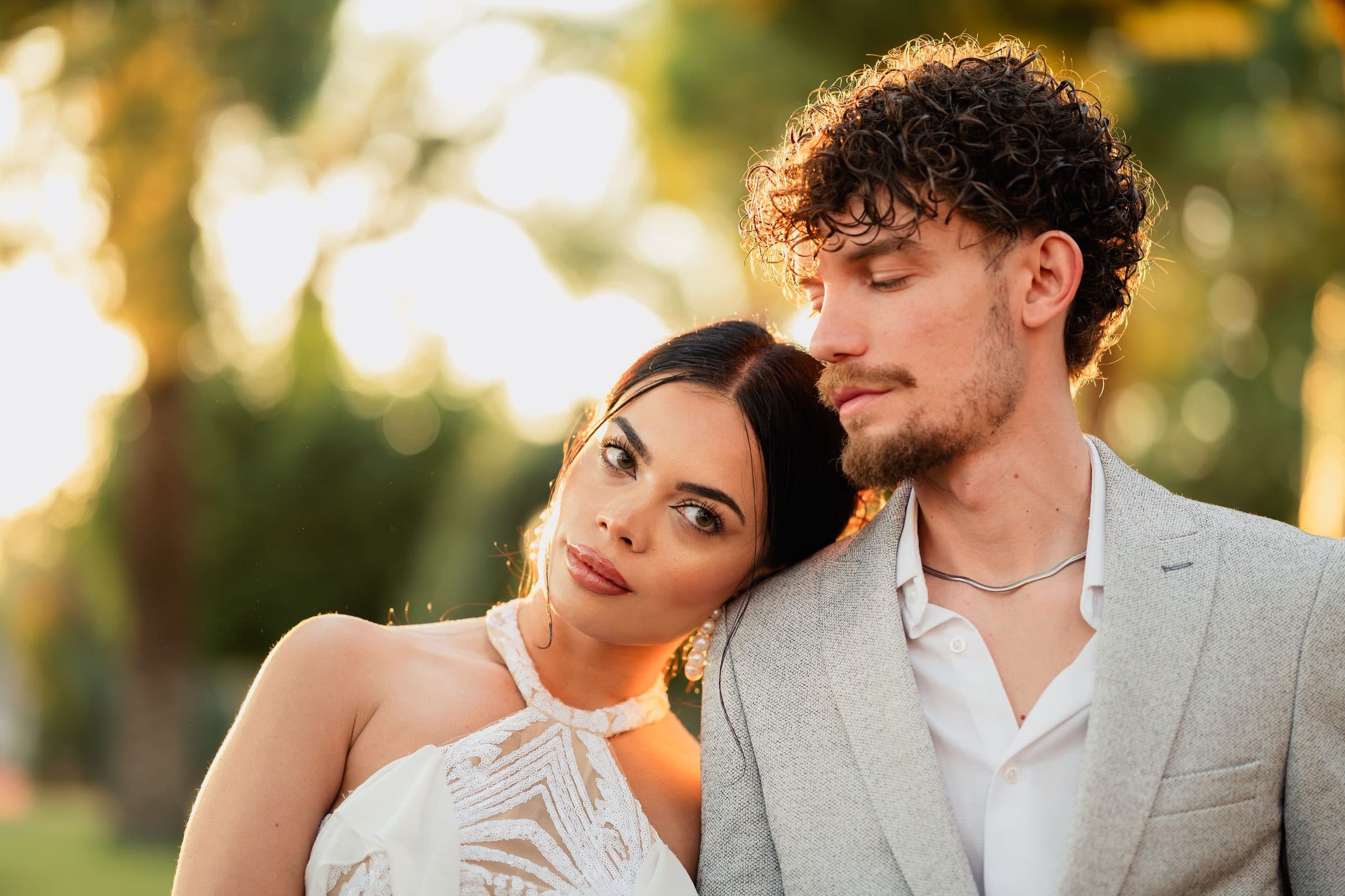 Woman with sleek dark hair in a white halter dress resting her head on the shoulder of a curly-haired man at golden hour