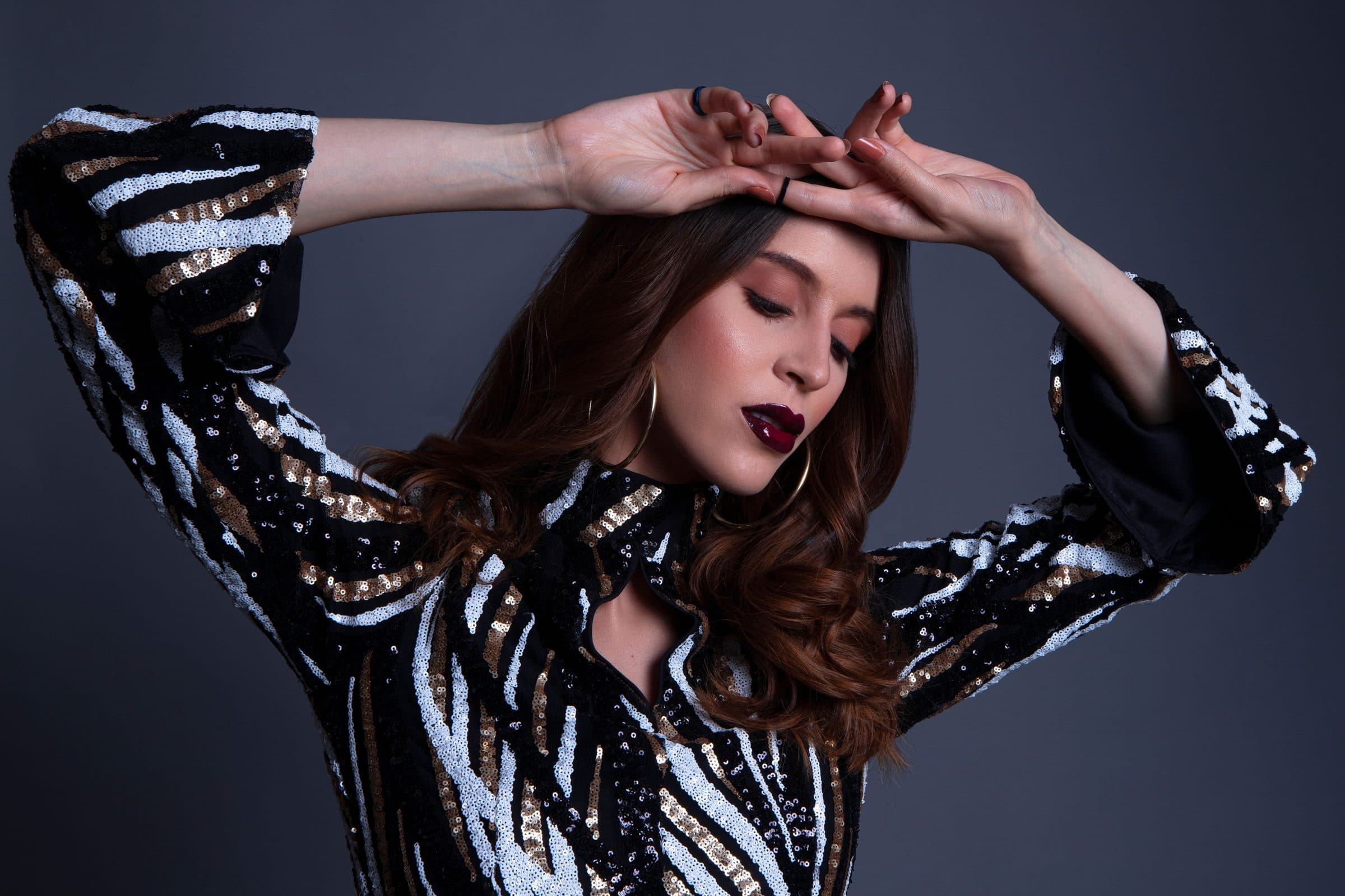 Woman with wavy auburn hair and dark lipstick in a sequin jacket, hands resting on her head