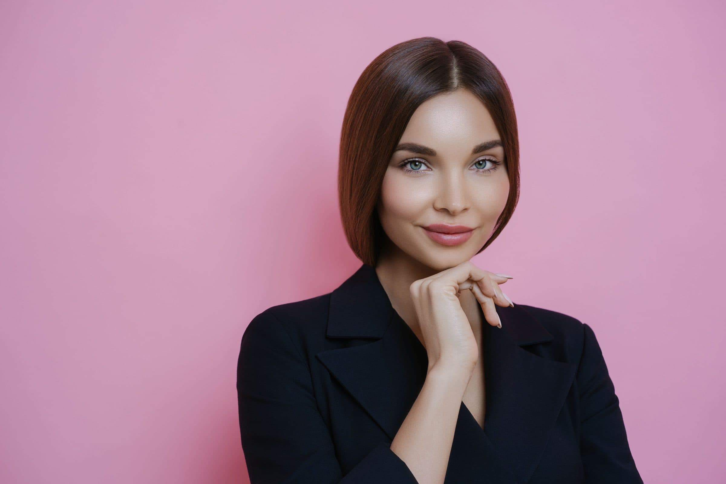 Smiling woman with a sleek brown bob and blazer resting her chin on her hand against a pink wall