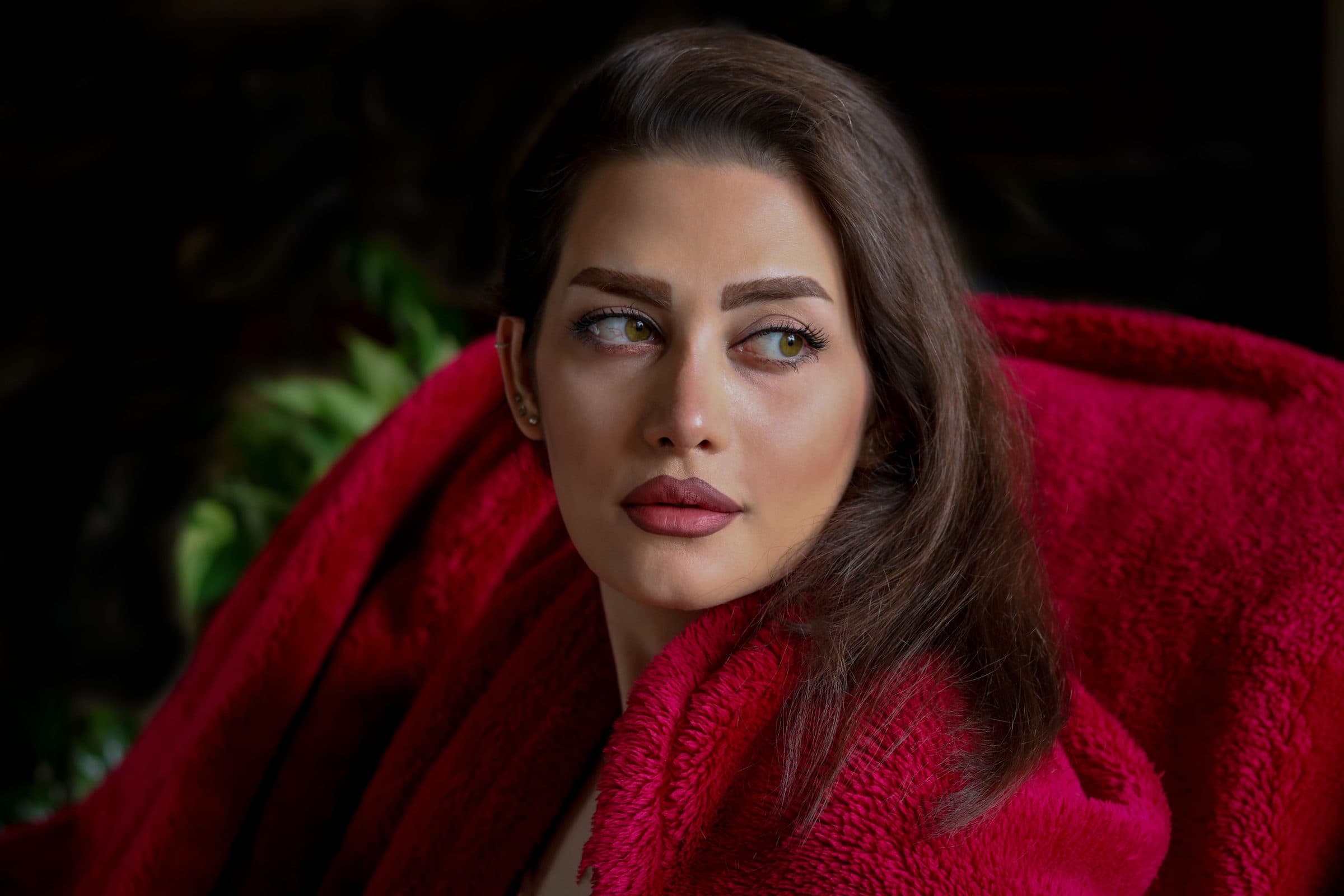 Woman with long brown hair wrapped in a red fluffy coat glancing to the side against a dark background
