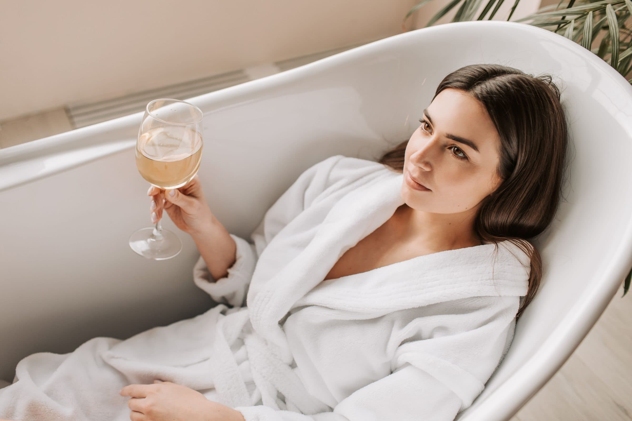 Woman with dark hair in a white robe reclining in a bathtub holding a glass of wine