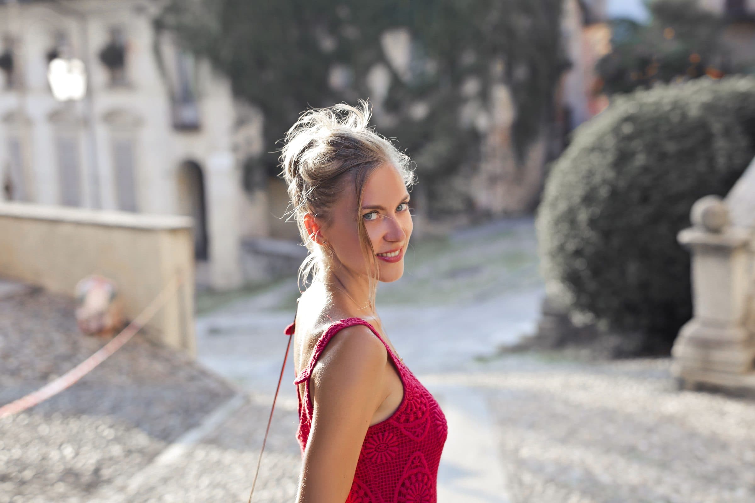Smiling blonde woman in a red lace dress looks back over her shoulder on a sunny cobblestone street