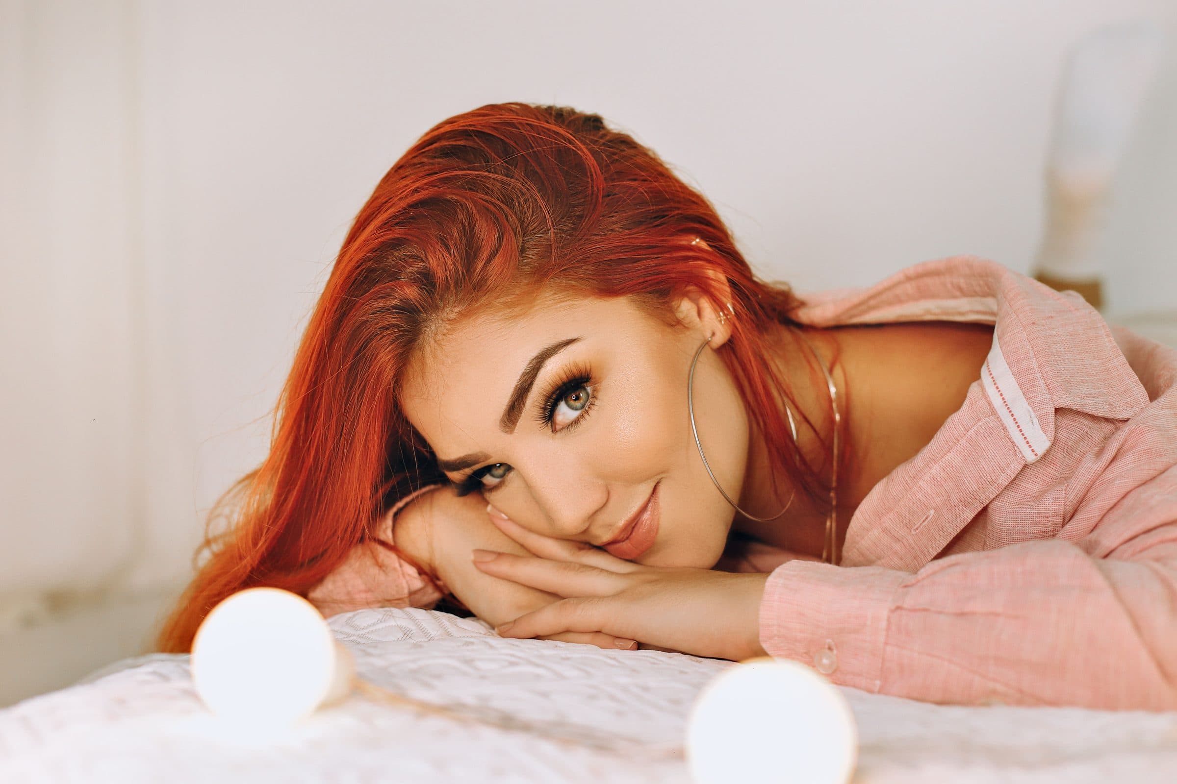 Woman with long red hair lying on a bed with her chin on her hands, smiling, in a pink shirt beside small globe lights
