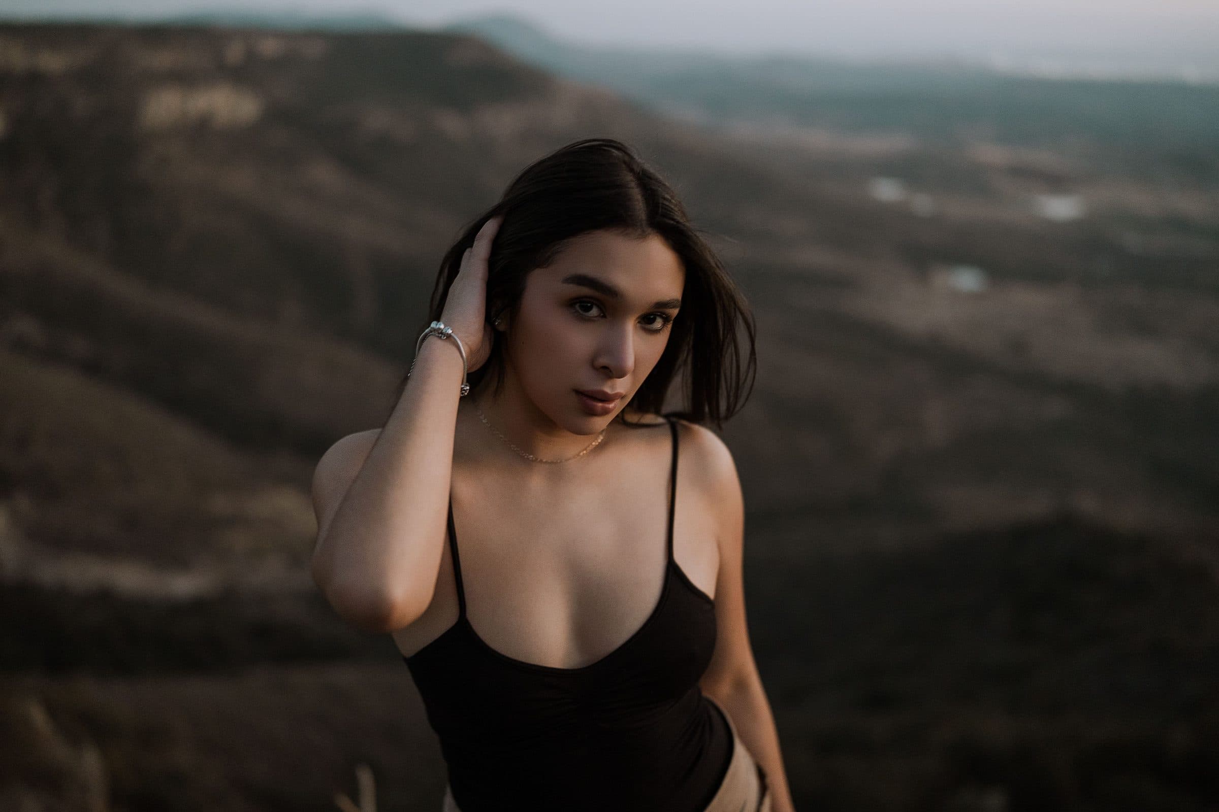 Woman with dark shoulder-length hair in a black bodysuit, hand in her hair, hills behind her
