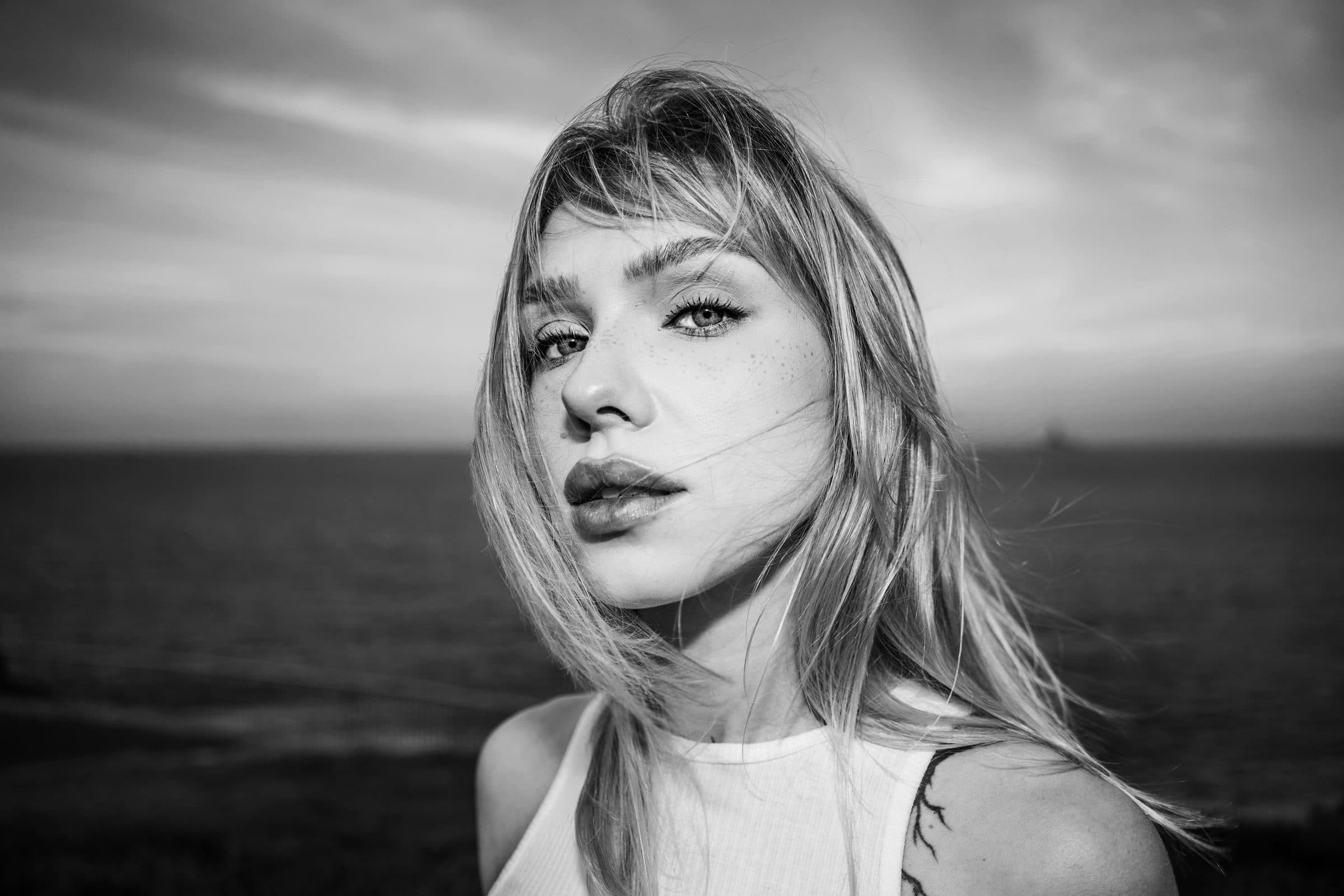 Black and white portrait of a woman with long hair and bangs in a white top by the sea