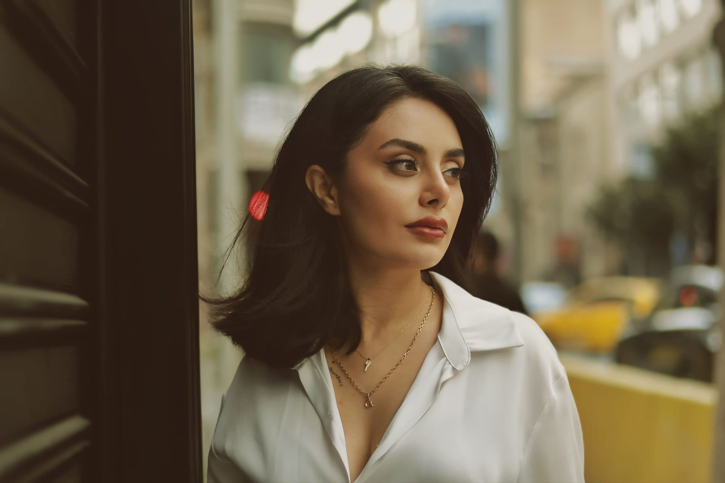Woman with shoulder-length dark hair in an open white shirt and gold necklaces looking down a city street