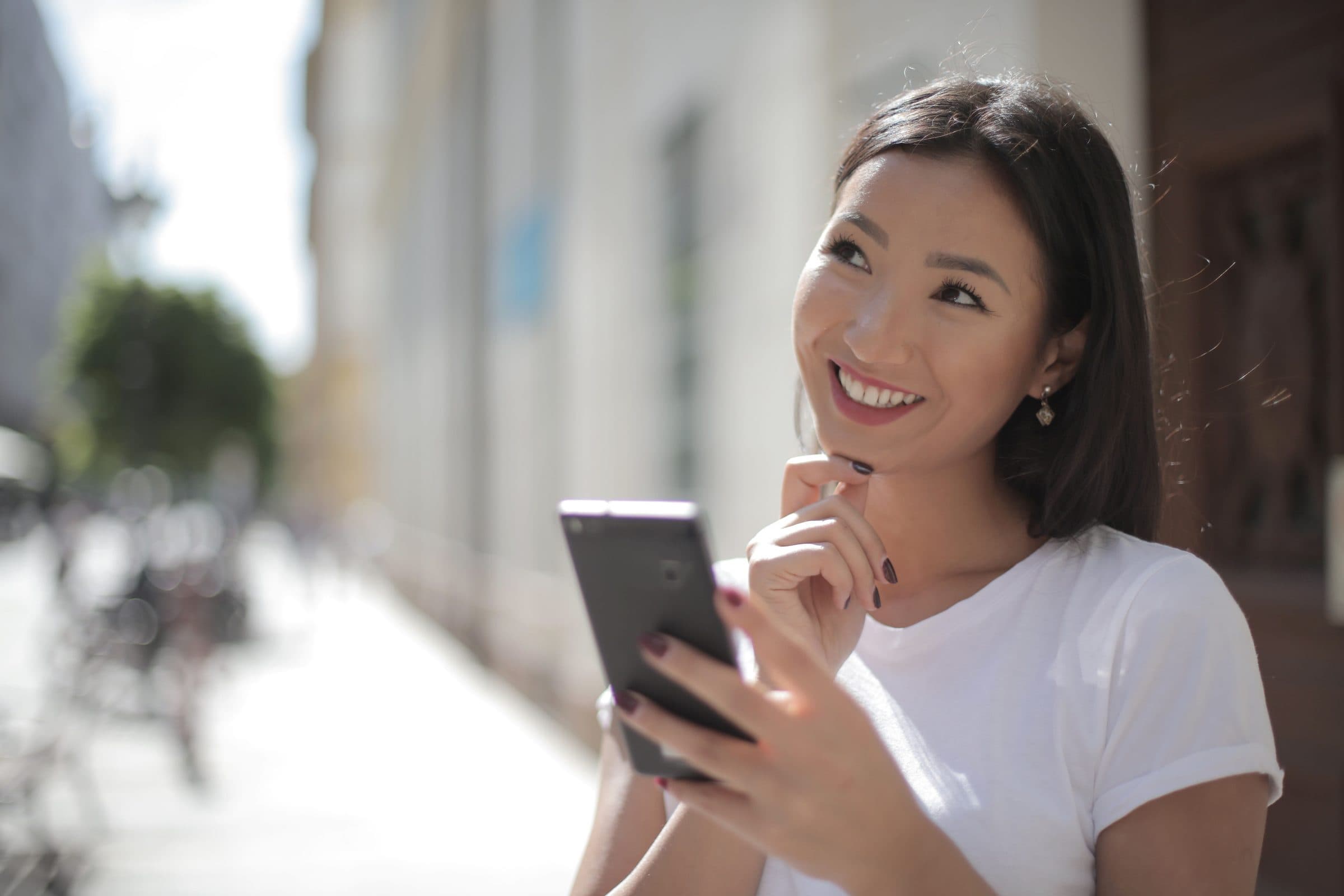 Smiling woman with shoulder-length dark hair in a white t-shirt holding a phone on a sunny street