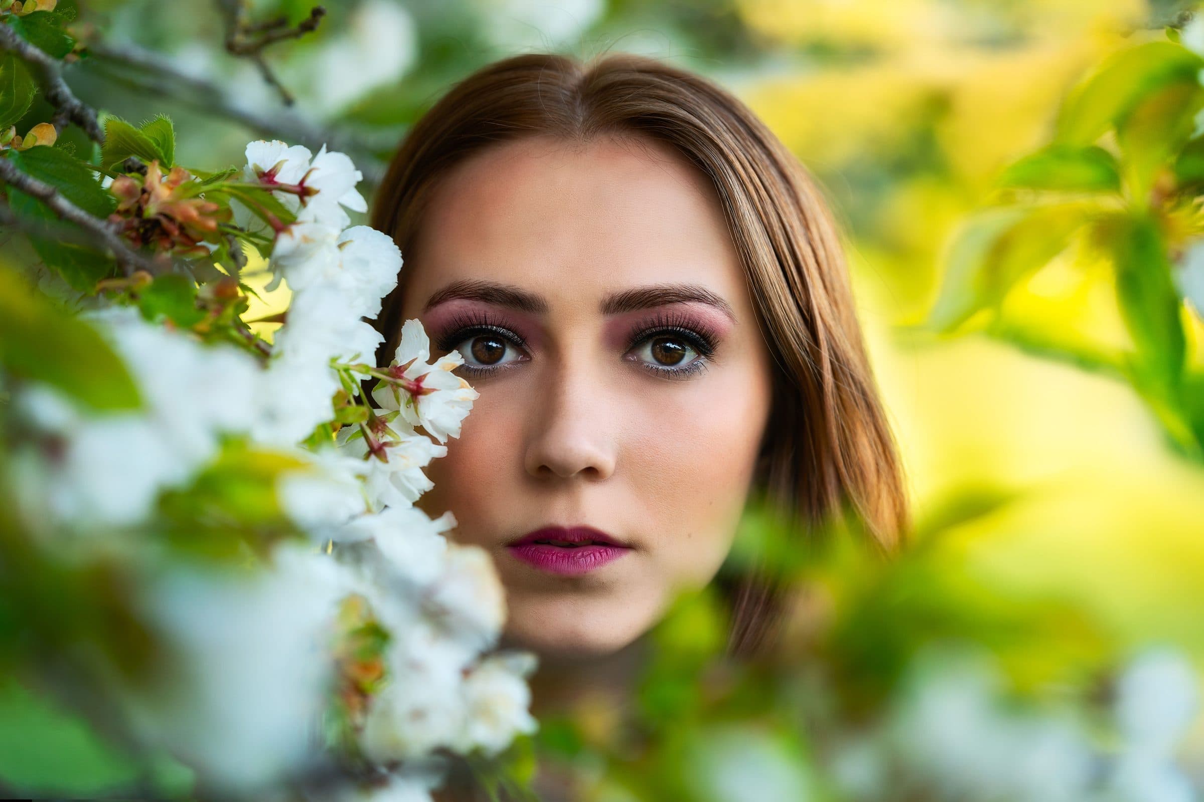 Woman with long light brown hair and bold eye makeup gazing at the camera beside sprays of white blossom