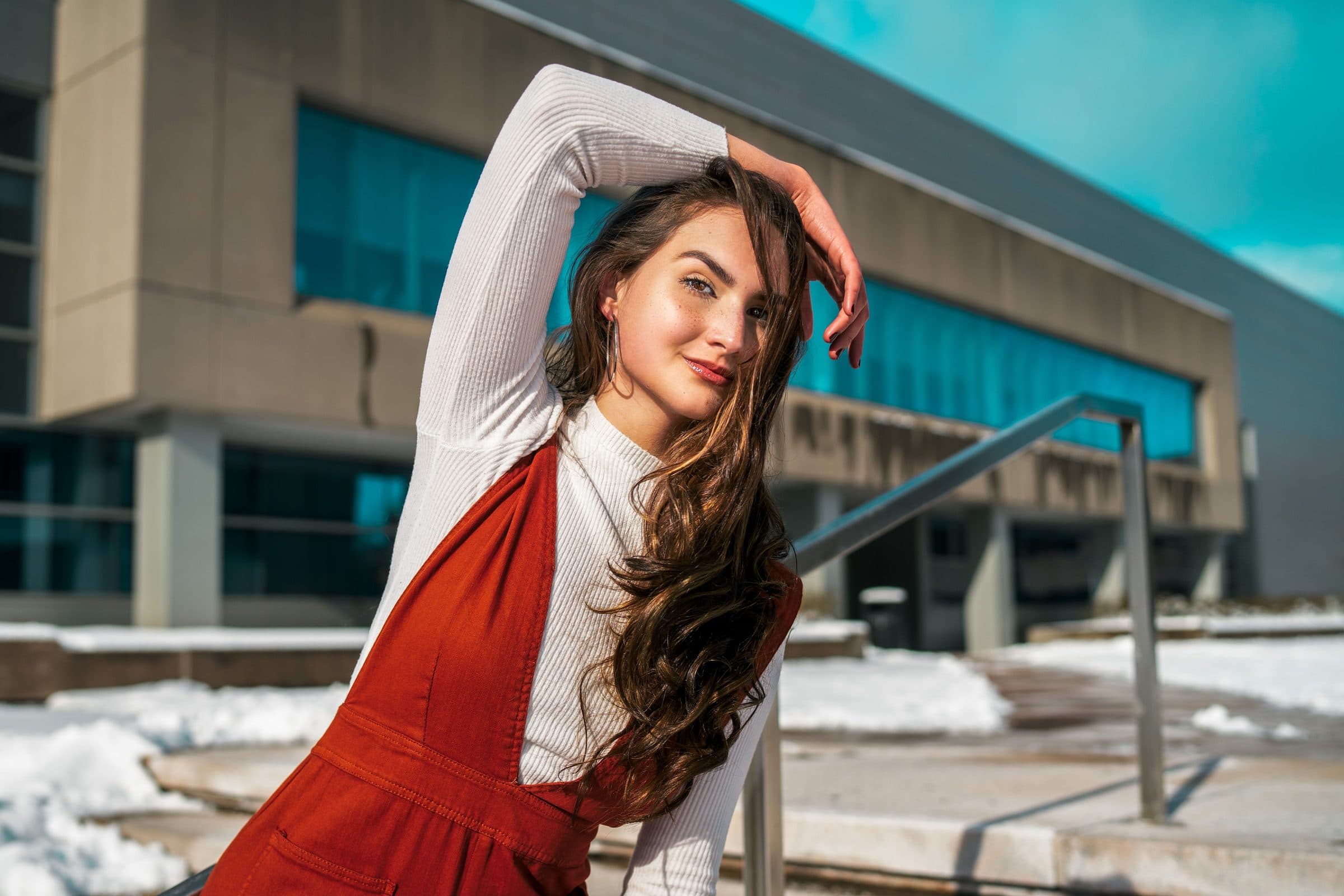 Woman with long brown hair in a red pinafore over a white top, arm raised, outside a modern building in snow