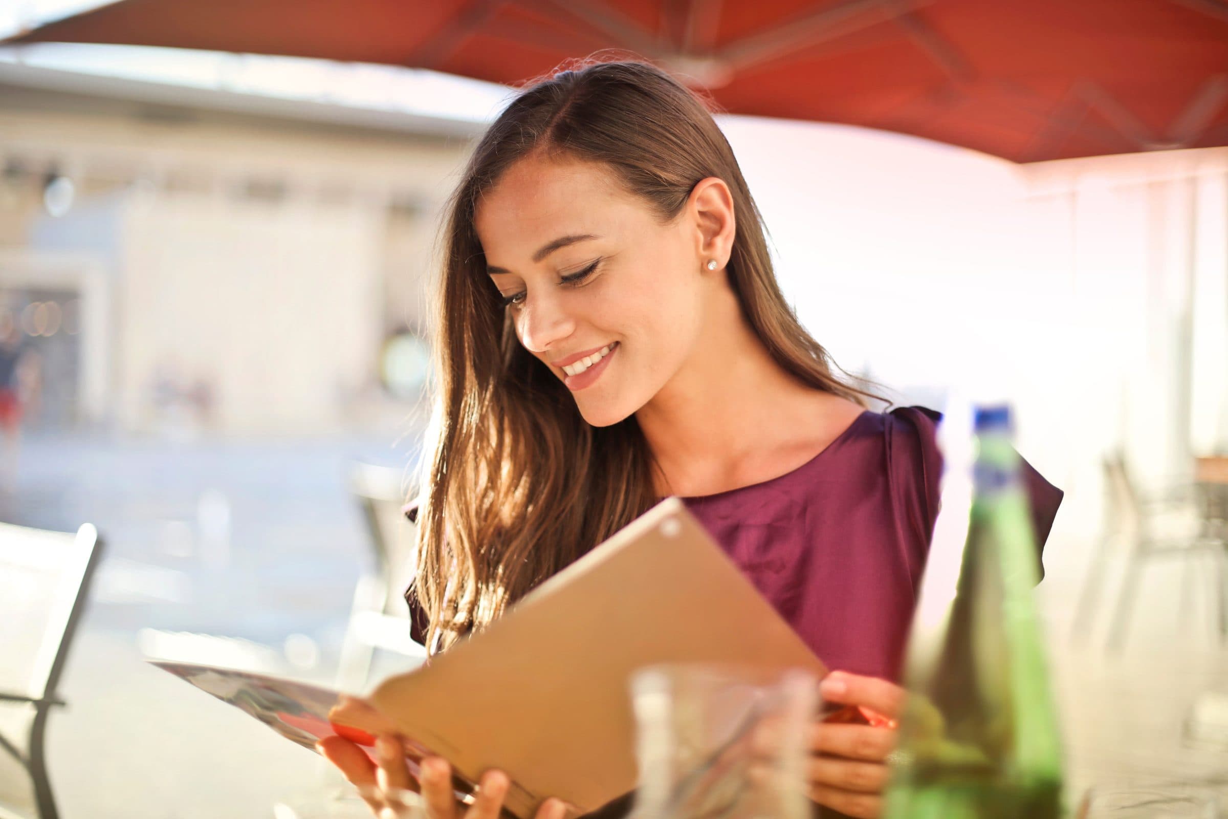 Smiling woman with long brown hair reading a menu at an outdoor cafe table under a red umbrella