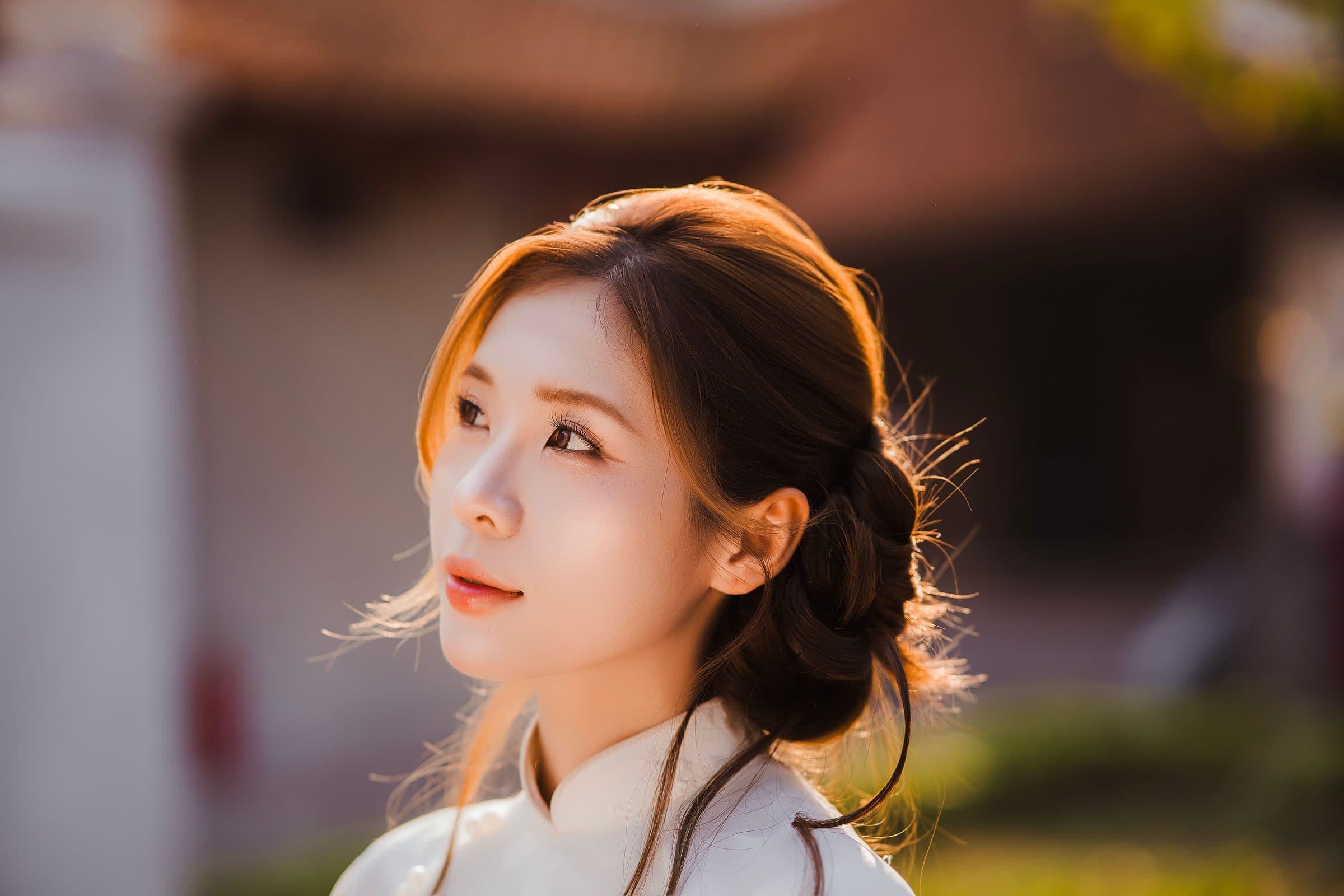Woman with dark hair in a low updo and a white high-collar top gazing upward in warm light
