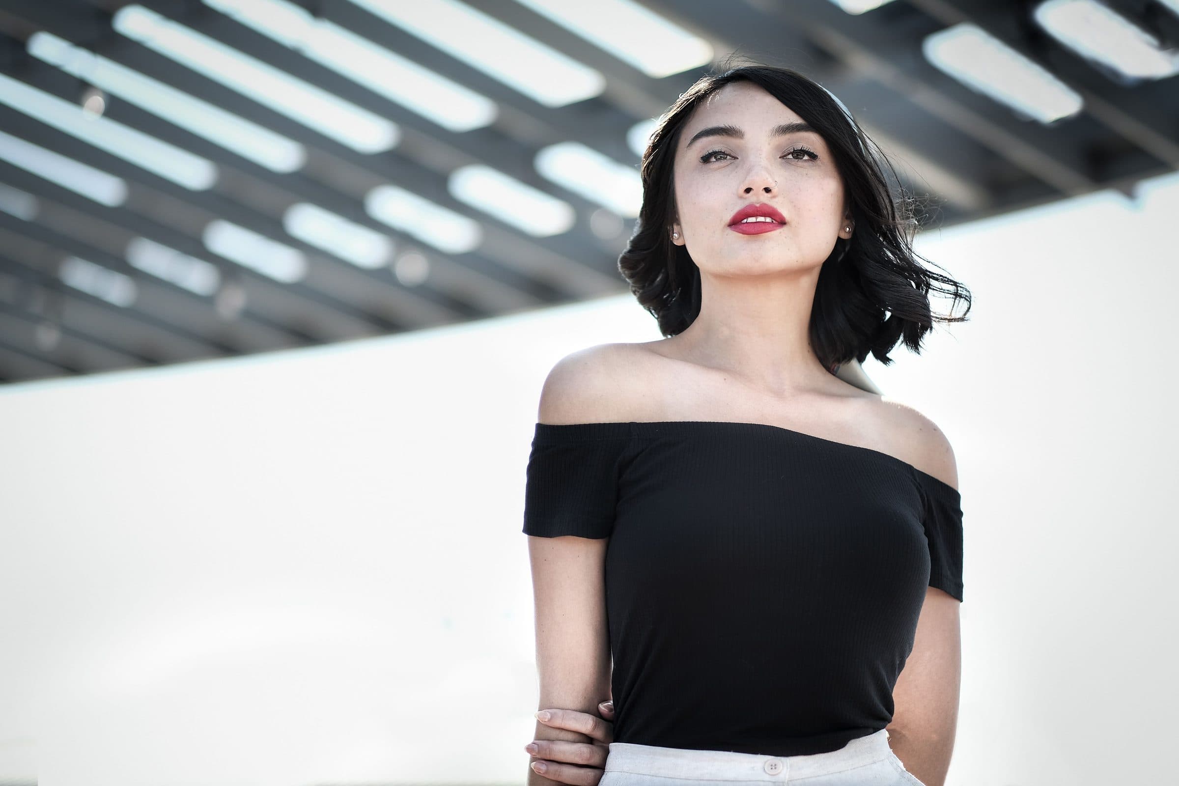 Woman with a black bob, red lips and an off-shoulder black top standing under a white pergola