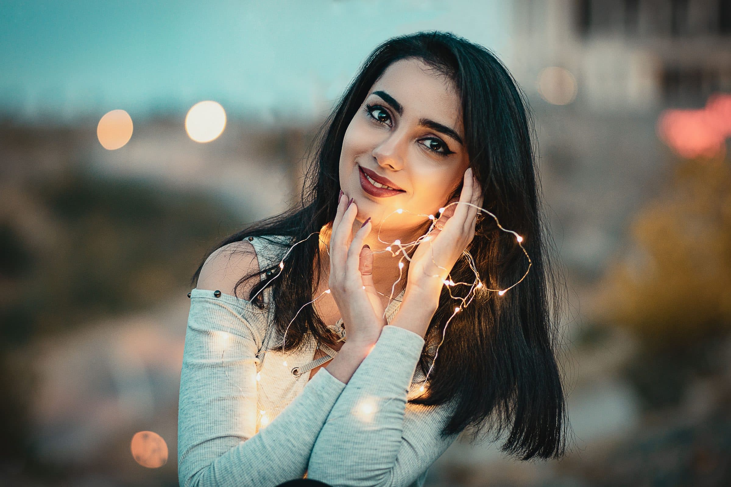 Smiling woman with long dark hair holding fairy lights near her face at dusk