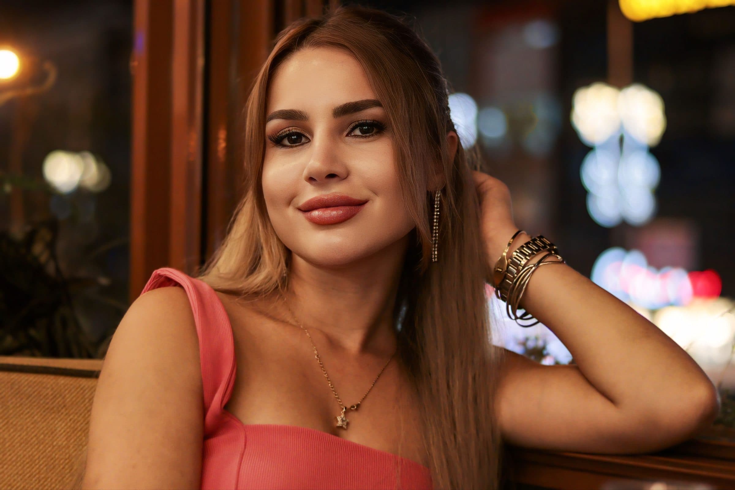 Smiling woman with long hair in a pink top sits by a window in a warmly lit restaurant at night