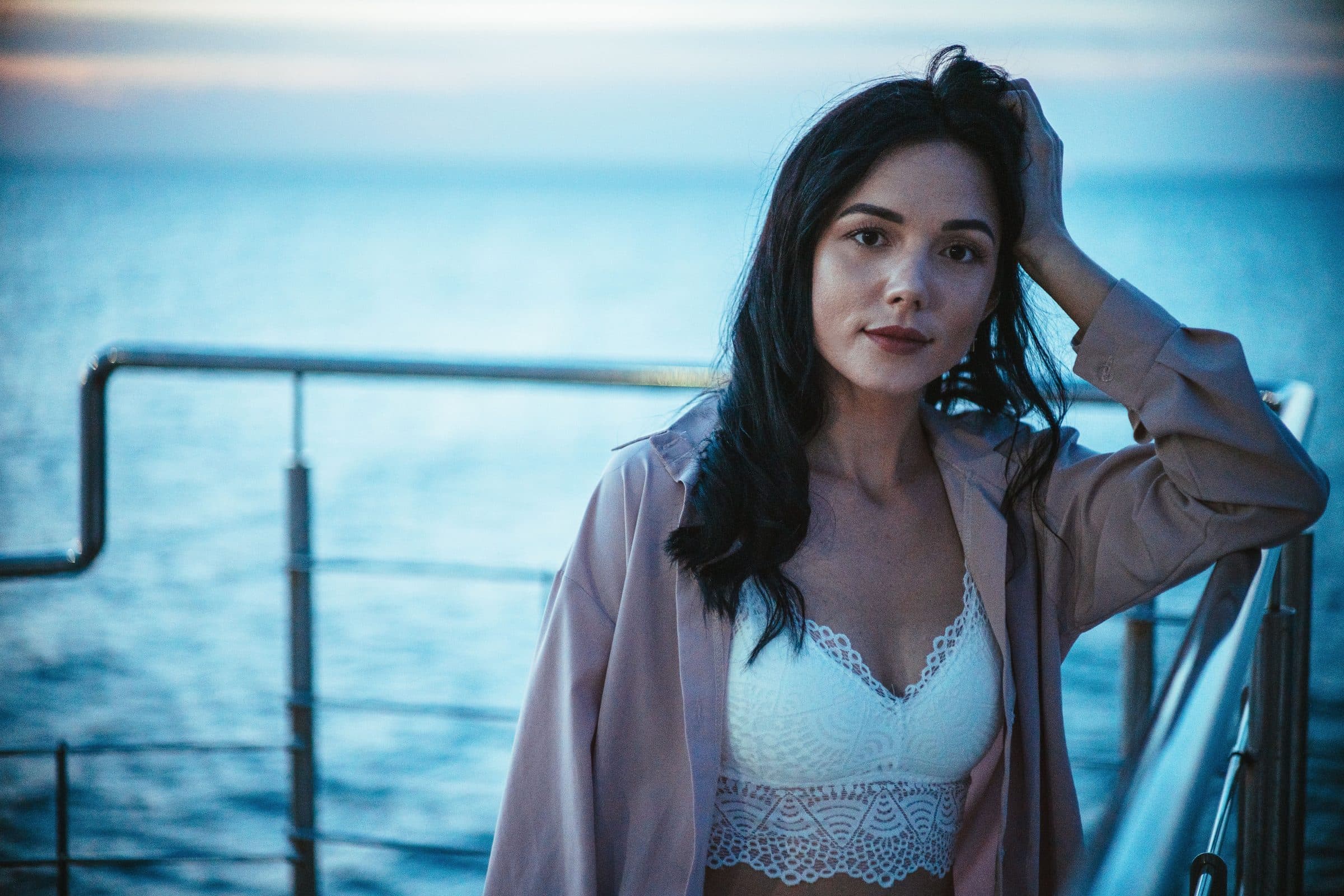 Woman with long dark hair and a hand in her hair standing at a boat railing over the sea at dusk