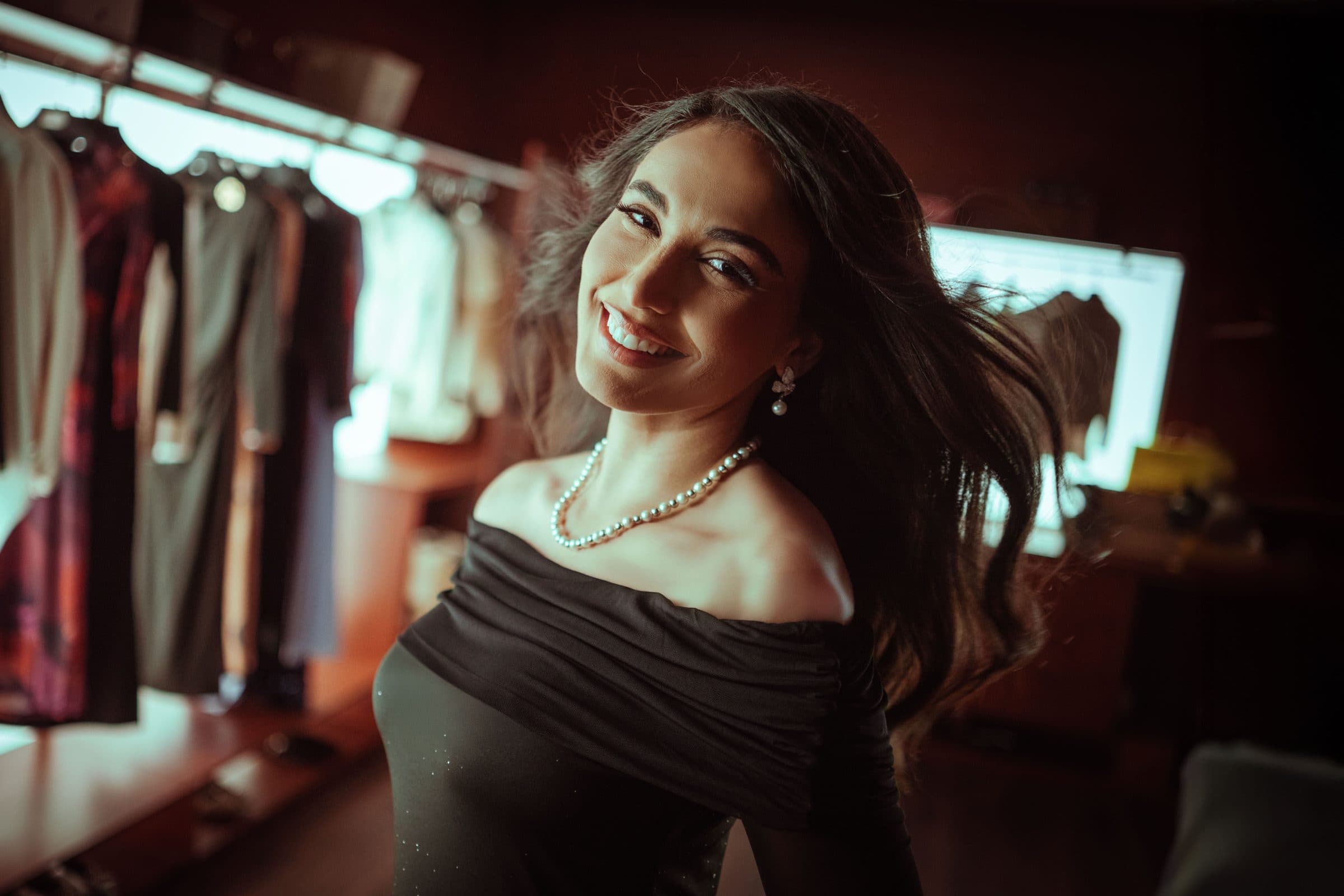 Smiling woman with long dark hair, an off-shoulder black top and pearls glancing back at the camera in a clothing boutique